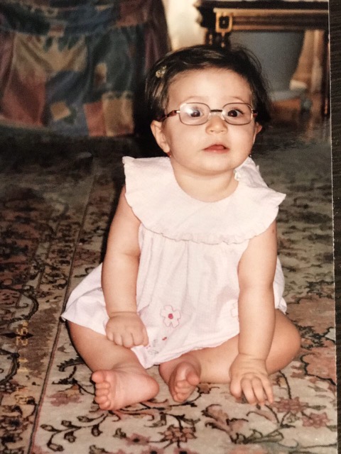 Romina as a baby wearing glasses and a pink dress